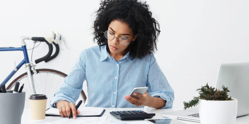 Business, career and job concept. Successful young dark-skinned female entrepreneur holding mobile phone, dialing number, looking at piece of paper, busy doing accounts, sitting at her workplace